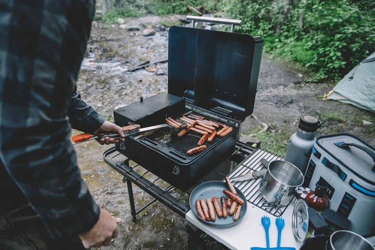 A person stands over a grill at a campsite cooking hot dogs